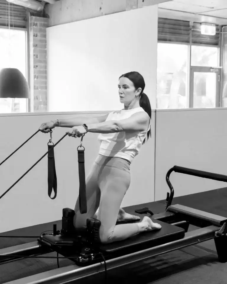 Woman kneeling on a Pilates reformer, performing an arm and back exercise with the straps